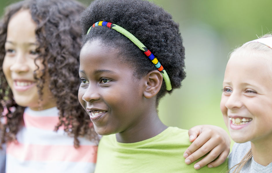 Three school-aged kids stand shoulder-to-shoulder smiling.  They are outside and not looking into the camera. 