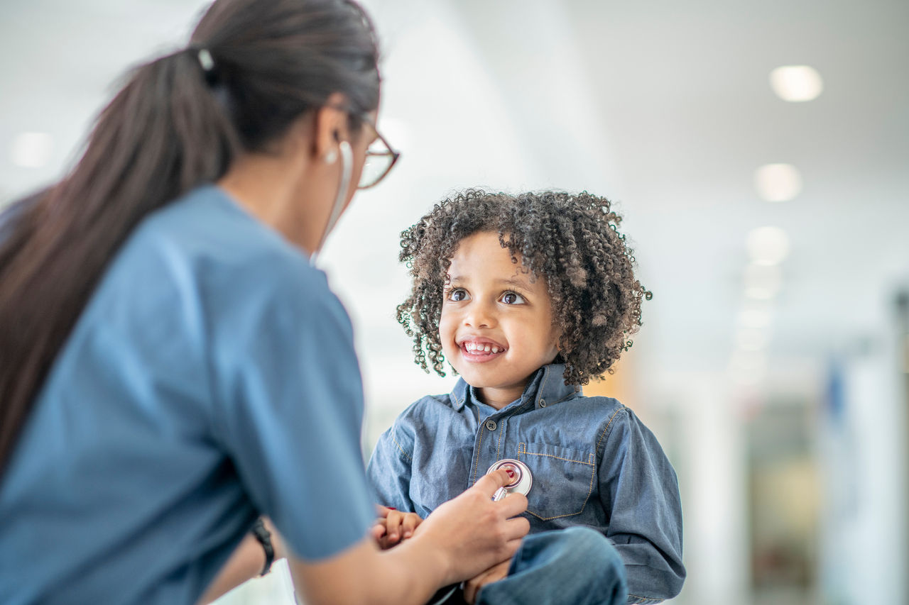 A black toddler boy sits on a table during a medical examination. A female medical professional is using a stethoscope to listen to his heartbeat. The child is smiling up at his doctor.