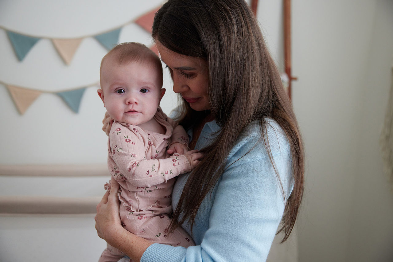 Person holding a baby dressed in a light pink outfit with a floral pattern, standing indoors near a wall decorated with triangular fabric bunting in muted colors.