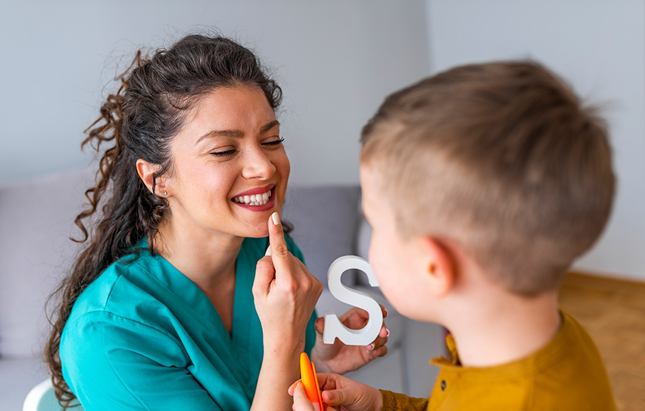 Young boy works with female speech therapist, she wears scrubs and holds the letter "S"